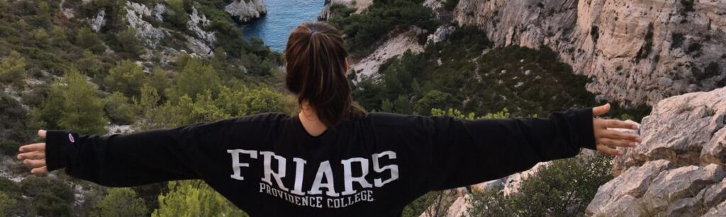 student looking out over a mountain cliff with her arms spread open wearing a shirt that says friars